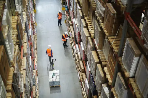 Warehouse staff moving a pallet between tall racks of stock.